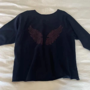 Svart offshoulder tröja med broderade vingar - Snygg svart tröja med offshoulder-design och broderade vingar på ryggen. Perfekt för en avslappnad stil med en unik touch. Passar bra till jeans eller kjol.