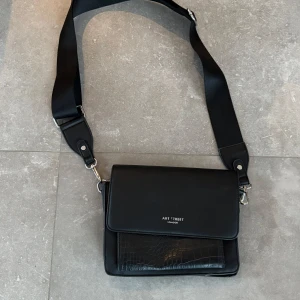 Svart axelväska från Art Treet - Snygg svart axelväska med justerbar rem och krokodilmönstrad detalj på framsidan. Väskan har en praktisk magnetknapp för enkel öppning och stängning. Perfekt för att bära dina dagliga nödvändigheter med stil.