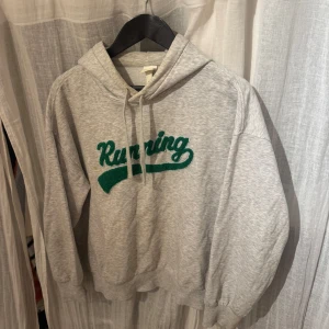 Grå hoodie med grön text - Säljer en grå hoodie med texten 'Running' i grönt på framsidan. Tröjan har en klassisk design med huva och dragsko samt långa ärmar. Perfekt för en avslappnad stil.
