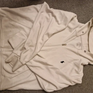 Vit hoodie från Polo Ralph Lauren - Säljer en stilren vit hoodie från Polo Ralph Lauren med dragkedja och det klassiska broderade logotypen på bröstet. Perfekt för en avslappnad look. Storlek M.