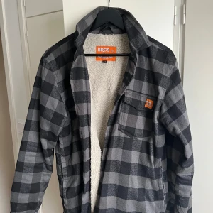 Fleecejacka från Bro’s workwear - Säljer en snygg svart och grå rutig jacka från Bros med mjukt fleecefoder. Jackan har långa ärmar och en praktisk bröstficka med knapp. Perfekt för kyliga dagar!