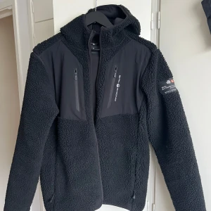 Svart teddyjacka Sail Racing - Säljer en svart teddyjacka från Sail Racing  med huva och dragkedja. Jackan har två bröstfickor med dragkedjor och en mjuk, fluffig yta. Perfekt för kyliga dagar. Köpt för 2399.