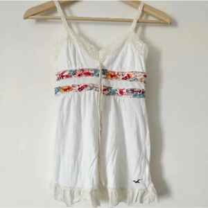 Vit linne med blommigt mönster från Hollister - Charmig vit linne/topp från Hollister med blommigt mönster i rött, blått och gult runt midjan. Klänningen har smala axelband och spetsdetaljer vid halsringningen. Den är kort och har volangdetaljer vid fållen. Perfekt för en somrig look! Storlek XS