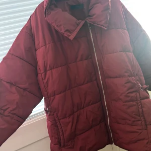 Vinröd pufferjacka från Ellos - Säljer en vinröd pufferjacka från Ellos med dragkedja och justerbart bälte i midjan. Jackan har långa ärmar och en hög krage för extra värme. Perfekt för kyliga dagar! Liten fläck (se bild 2), går ev bort i tvätten.