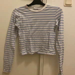 Randig långärmad topp från H&M - Säljer en randig långärmad topp från H&M i ljusblått och vitt. Perfekt för en avslappnad stil. Toppen har en rund halsringning och är gjord i ett mjukt material.