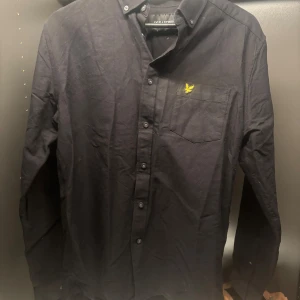 Svart skjorta från Lyle & Scott - Snygg svart skjorta från Lyle & Scott med klassisk design. Skjortan har långa ärmar och en bröstficka med en gul broderad logotyp. Perfekt för en stilren look.