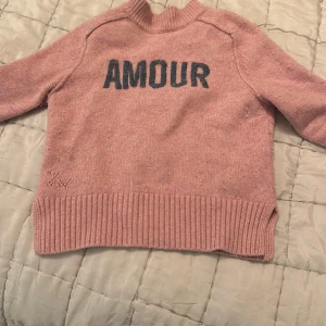 Rosa stickad tröja med text - Säljer en rosa stickad tröja med texten 'AMOUR' i svart på framsidan. Tröjan har långa ärmar och en ribbad kant vid ärmslut och nederkant. Perfekt för en mysig dag.