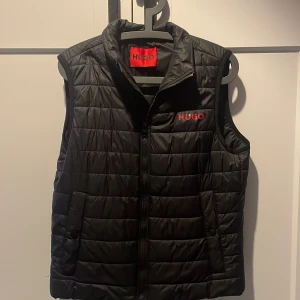 Svart quiltad väst från Hugo - Snygg svart quiltad väst från Hugo med dragkedja framtill. Perfekt för lager-på-lager stil och håller dig varm under kyliga dagar. Märkeslogga i rött på bröstet ger en stilren touch.