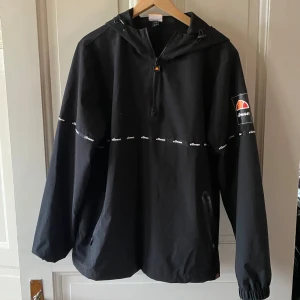 Vindjacka från Ellesse - Säljer en svart Anorak från Ellesse med huva och dragkedja. Jackan har ett tryckt Ellesse-märke på ärmen och ett band med logotypen runt bröstet. Perfekt för blåsiga dagar.💯