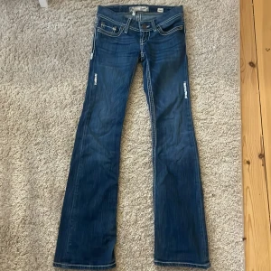 Blå bootcut jeans från BKE - Snygga blå bootcut jeans från BKE med dekorativa sömmar på bakfickorna. Jeansen har låg midja och klassisk femficksdesign med knappar och dragkedja framtill. Perfekta för en avslappnad stil.
