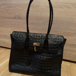 Svart krokodilpräglad handväska - Elegant svart handväska med krokodilpräglat mönster och guldfärgad låsdetalj. Väskan har två handtag och en stilren design som passar perfekt för en chic look.