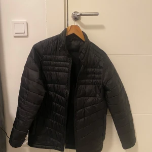 Svart pufferjacka från Jack & Jones - Säljer en svart pufferjacka från Jack & Jones. Jackan har en dragkedja framtill och är quiltad för extra värme. Perfekt för kyligare dagar med sin stilrena design och bekväma passform. Perfekt för hösten och våren. Storlek 164