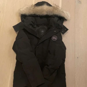 Svart dunjacka från Canada Goose - Säljer en svart dunjacka från Canada Goose med pälskantad huva. Jackan har en klassisk design med långärmade ärmar och en broderad logotyp på ärmen. Perfekt för kalla vinterdagar.