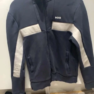 Mörkblå kofta från Boss - Snygg mörkblå kofta från Boss med vita detaljer och dragkedja. Tröjan har en sportig look med ribbade partier på ärmarna och sidorna. Perfekt för en stilren och avslappnad stil.