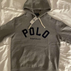 Grå hoodie från Ralph Lauren - Säljer en stilren grå hoodie från Ralph Lauren med stor POLO-logga i svart på bröstet. Tröjan har en klassisk känguruficka och justerbar huva med vita snören. Perfekt för en avslappnad stil.