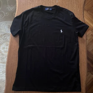 Svart t-shirt från Polo Ralph Lauren - Snygg svart t-shirt från Polo Ralph Lauren med klassisk passform. Den har en liten broderad logga i vitt på bröstet. Perfekt för en stilren look.