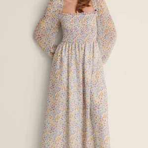 Blommig långklänning med smockdetaljer - Charmig, slutsåld långklänning med blommigt mönster i pastellfärger. Klänningen har smockdetaljer över bysten och långa, transparenta ärmar. Den perfekta klänningen inför midsommar. Helt ny och oanvänd med prislapp kvar. Enbart testad. 