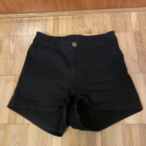 Svarta shorts med hög midja - Snygga svarta shorts med hög midja och knappstängning fram. De har två bakfickor och är perfekta för en stilren look. Passar bra till både vardag och festliga tillfällen.