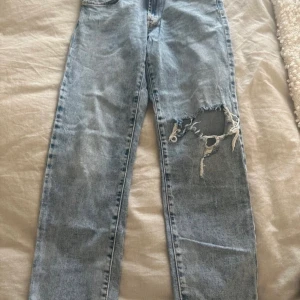 Ljusa jeans från Zara - Säljer ett par ljusa jeans från Zara med slitna detaljer på ena benet. De har en klassisk femficksdesign och knappgylf. Perfekta för en avslappnad stil.