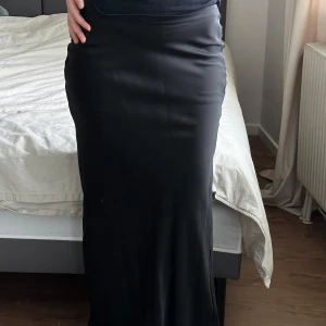 Svart långkjol i satin - Elegant svart långkjol i satin med en slät och glansig yta. Kjolen är figurnära upptill och faller vackert ner mot golvet. Storlek 36! Använd 2-3 ggr, fortfarande i jättefint skick! Jag är 165cm för referens!