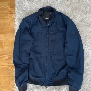 Mörkblå jacka från J.Lindeberg - Snygg mörkblå jacka från J.Lindeberg med dragkedja och ribbade detaljer vid ärmslut och nederkant. Skick 9/10, perfekt för en stilren look. Jackan har en diskret bröstficka med dragkedja och en liten logotyp på ärmen. Storlek M, passar någon 175/165 cm och vikt mellan 55/65 kg 😇