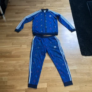 Unikt träningsset från Adidas - Säljer ett blått träningsset från Adidas med vita ränder längs ärmar och ben. Setet har ett unikt mönster. Perfekt för sportiga aktiviteter eller en avslappnad stil.  Settet är i mycket bra skick och är sällan använt. Tröjan är i storlek 176cm och byxorna är i storlek 164cm. Tveka inte att höra av er!🙂