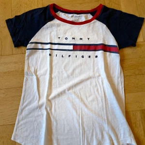 Vit och marinblå t-shirt från Tommy Hilfiger - Snygg vit t-shirt med marinblå ärmar och röd krage från Tommy Hilfiger. T-shirten har ett tryck med märkesnamnet och en röd och blå rand över bröstet. Perfekt för en avslappnad stil. Stl xxs.