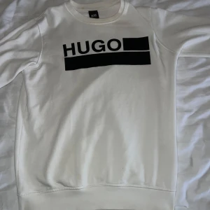 Vit sweatshirt från Hugo Boss - Säljer en stilren vit sweatshirt från Hugo Boss med svart logotyp på bröstet. Tröjan har rund hals och långa ärmar, perfekt för en avslappnad look. Passar bra till både jeans och joggers.