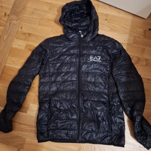Svart pufferjacka från Emporio Armani - Säljer en helt nytt svart pufferjacka från Emporio Armani med huva och dragkedja. Jackan är quiltad och har en stilren design med EA7-logga på bröstet. Perfekt för kyliga dagar och lätt att packa ner i medföljande påse. Anledning för att jag säljer den är att den är för liten för mig.