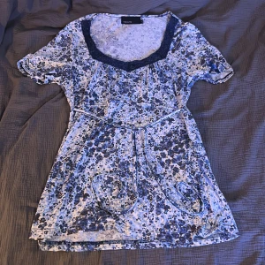 Blommig blus från KappAhl - Säljer en blommig blus från KappAhl i storlek M.  Blusen har korta ärmar och en v-ringad spetsdetalj vid halsen. Den är i en ljus färg med ljus och mörkblåa blommor samt ett knytband i midjan. Väldigt avlång i passform 