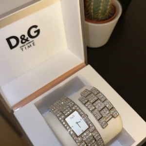 Glittrig klocka från D&G - Snygg och glittrig klocka från D&G med ett fyrkantigt ur och gnistrande stenar på armbandet. Modell : ”C’est Chic” . Klockan har en elegant design med en silverfärgad urtavla och tydliga visare. Perfekt för att ge en touch av glamour till din stil. Äkta klocka - nytt pris över 4000 kr. Vid snabb affär går det att få billigare. 