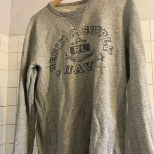 Grå tröja från Denim & Supply Ralph Lauren - Säljer en grå tröja från Denim & Supply Ralph Lauren med ett tryck på framsidan som inkluderar ett ankare och texten 'NAVY'. Tröjan har långa ärmar och en ribbad krage. Perfekt för en avslappnad stil.