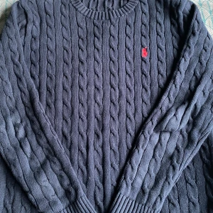 Mörkblå kabelstickad tröja från Ralph Lauren - Säljer en stilren mörkblå kabelstickad tröja från Ralph Lauren med det klassiska röda logotypbroderiet på bröstet. Tröjan har långa ärmar och ribbstickade muddar vid ärmslut och nederkant. Perfekt för kyliga dagar!