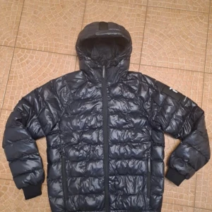 Svart dunjacka från Peak Performance - Snygg svart dunjacka från Peak Performance med huva och dragkedja. Perfekt för kyliga dagar. Jackan har en stilren design med märkets logga på ärmen.