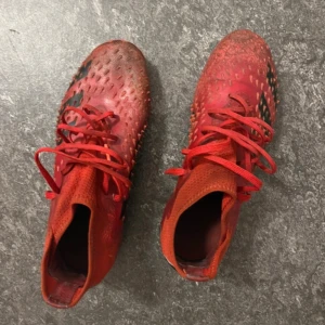 Röda Adidas fotbollsskor - Säljer ett par röda Adidas fotbollsskor med svarta detaljer och snörning som finns i bra skick. Skorna har en unik struktur på ovansidan och är designade för optimalt grepp på planen. Perfekta för fotbollsspelare som vill ha stil och funktion.