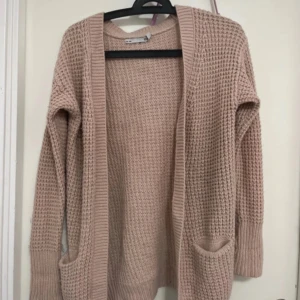 Beige stickad kofta från 157 - Mysig beige stickad kofta från 157 med långa ärmar och praktiska fickor framtill. Perfekt för kyliga dagar och ger en avslappnad look. Koftan har en öppen design utan knappar.