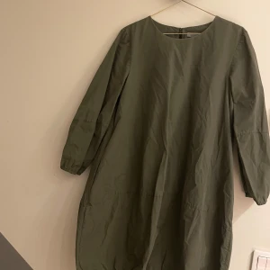 Olivgrön klänning från COS - Snygg olivgrön klänning från COS med lång ärm och rund hals. Tillverkad i ett lätt och bekvämt material, perfekt för en stilren look. Klänningen har en diskret dragkedja i ryggen för enkel påklädning.