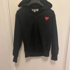 Svart hoodie från Comme des Garçons - Säljer en stilren svart hoodie från Comme des Garçons med det ikoniska röda hjärtat på bröstet. Tröjan har dragkedja och huva, perfekt för en casual look. Passar utmärkt till både jeans och joggers.