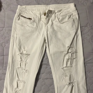 Vita jeans med slitningar och dragkedjor lågmidjade - Snygga vita jeans från Cobrex med slitningar på framsidan och dragkedjor på bakfickorna. De har en knappstängning och bälteshällor. Perfekta för en trendig look!