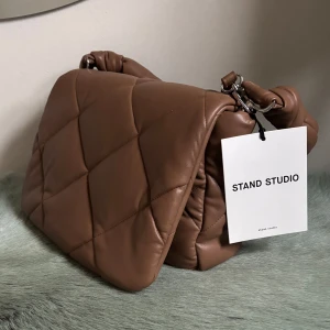 Brun quiltad bag från Stand Studio - Snygg brun quiltad väska från Stand Studio med en mjuk och vadderad design. Väskan har en praktisk dragkedja och en stilren look. Perfekt för att ge din outfit en trendig touch. Pris kan diskuteras 
