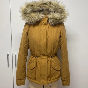 Gul parkajacka med fuskpäls - Snygg gul parkajacka med stor huva kantad av fuskpäls. Jackan har en dragsko i midjan för justerbar passform och praktiska fickor framtill. Perfekt för kyliga dagar.