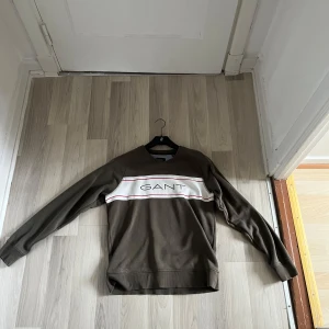 Brun tröja från GANT - Snygg brun tröja från GANT med vit och röd rand över bröstet och GANT-logga. Perfekt för en avslappnad stil. Långärmad och bekväm passform.