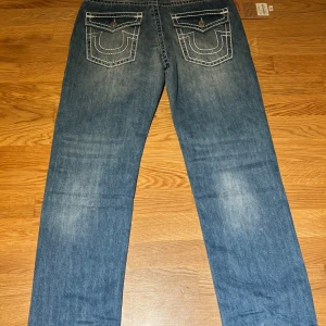 True Religion - Snygga blå jeans från True Religion med kontrasterande vit söm och klassisk femficksdesign. Byxorna har en straight passform och är dekorerade med knappar och broderade detaljer på bakfickorna.