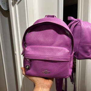 Lila ryggsäck från Coach - Snygg lila ryggsäck från Coach i skinn med dragkedja. Perfekt för att bära dina dagliga nödvändigheter med stil. Den har justerbara axelremmar och en praktisk ytterficka med dragkedja.