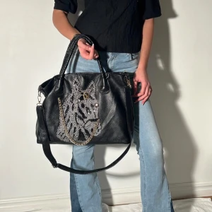 Svart axelväska med zebramönster - Snygg svart axelväska i skinn med ett iögonfallande zebramönster i paljetter på framsidan. Väskan har både handtag och en justerbar axelrem för flexibel användning. Perfekt för att ge din outfit en unik touch.