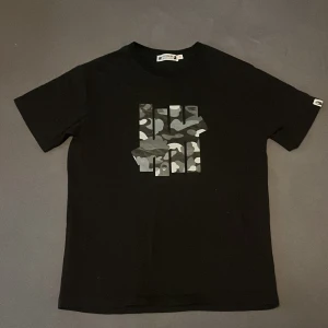 Svart t-shirt från Undefeated x A Bathing Ape - Snygg svart t-shirt med camouflagemönster och tryck från Undefeated x A Bathing Ape. T-shirten har korta ärmar och en normal passform. Perfekt för streetwear-entusiaster! Om minsta fundering eller fråga, är det bara kontakta. (Tar emot alla möjliga byten) byter hälst. Pris kan diskuteras.