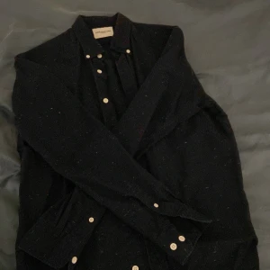 Svart skjorta från These Glory Days - Snygg svart skjorta från These Glory Days med vita knappar. Perfekt för en stilren look. Skjortan har långa ärmar och en klassisk krage. Använt kanske 2 gånger inga fläckar eller nått.