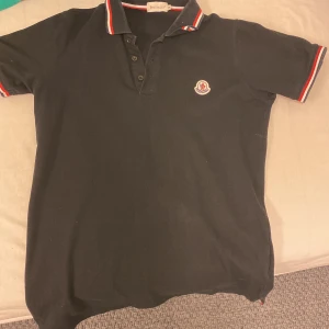 Svart pikétröja från Moncler - Snygg svart pikétröja från Moncler med klassisk krage och röd-vita detaljer. Tröjan har korta ärmar och en broderad logotyp på bröstet. Ingen skador eller nåt bra skick. Priset diskuteras för snabb affär
