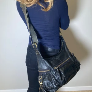 Svart axelväska med nitar - Snygg svart axelväska i skinn med coola nitar och dragkedjedetaljer. Perfekt för att ge din outfit en edgy touch. Väskan har en justerbar axelrem och flera praktiska fack för enkel organisering.