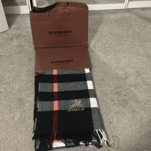 Rutig halsduk från Burberry - Snygg rutig halsduk från Burberry i svart, grått, vitt och rött. Den har ett klassiskt rutmönster och fransar i ändarna. Perfekt för att hålla dig varm och stilren under kyliga dagar.